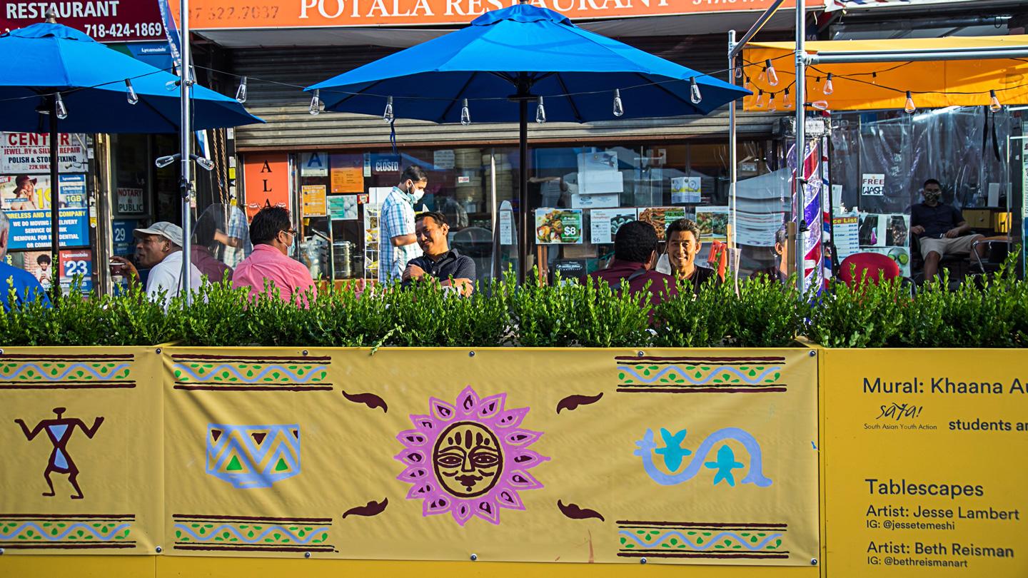 View of murals along street side dining with planters and umbrellas at DineOut Jackson Heights