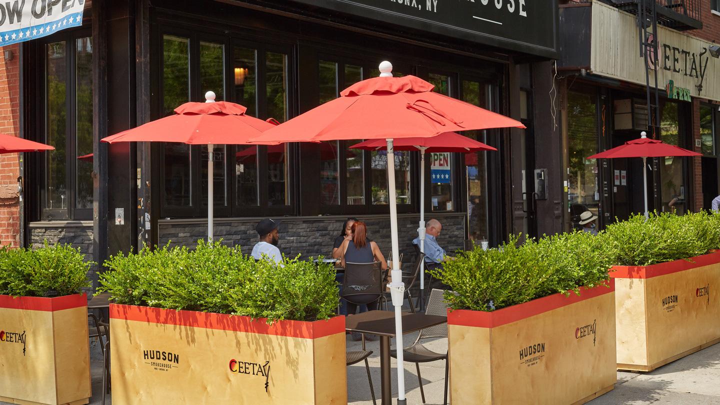 Umbrellas and sidewalk seating surrounded by planters outside of BBQ restaurant in Bronx